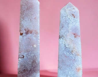 May include: Two light purple crystal points with a rough, textured surface. The crystals are standing upright on a pink background.