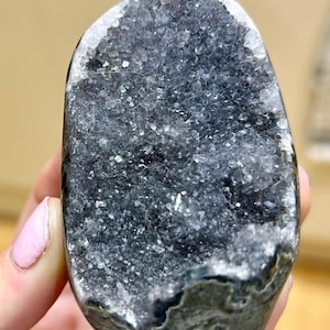 May include: A dark gray, naturally formed geode with a crystal-lined interior. The geode is smooth and rounded, with a slightly irregular shape.