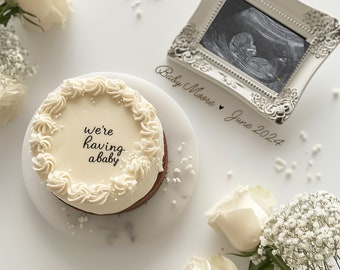 May include: A white cake with a white frosting and the words "we're having a baby" written in black. The cake is on a white plate with white flowers. There is a white frame with a black and white ultrasound image inside. The frame is on a white surface with the words "Baby Moore June 2024" written in black.