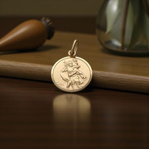May include: A gold pendant depicting Saint Christopher carrying a child. The circular pendant has a loop for a chain. The background includes a wooden surface and a blurred decorative object.