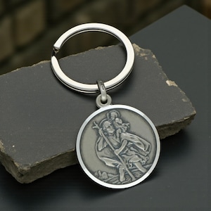 May include: A silver-colored Saint Christopher keychain. The round pendant features a detailed relief of Saint Christopher carrying a child, with a staff in hand. The keychain has a split ring for keys.
