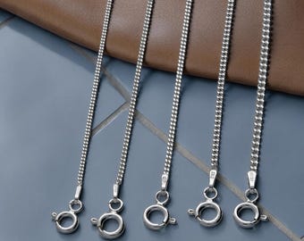 May include: Five silver curb chains of varying thicknesses, from 1mm to 2.1mm, are shown. Each chain has a clasp at the bottom. The chains are set against a brown leather surface and a blue tiled background.