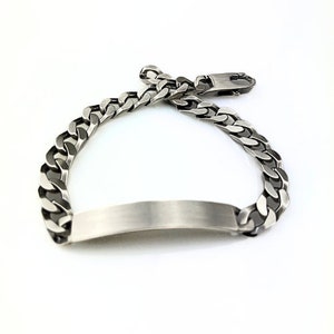 Könnte beinhalten: Ein silberfarbenes Panzerkettenarmband mit rechteckigem Namensschild. Das Armband verfügt über einen sicheren Verschluss und wird auf weißem Hintergrund präsentiert. Die Kettenglieder sind dick und flach.