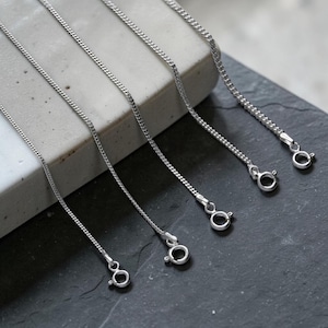 May include: Close-up of several silver-colored chain necklaces with lobster claw clasps. The chains are displayed on a gray and black surface. The chains have a delicate, textured design.