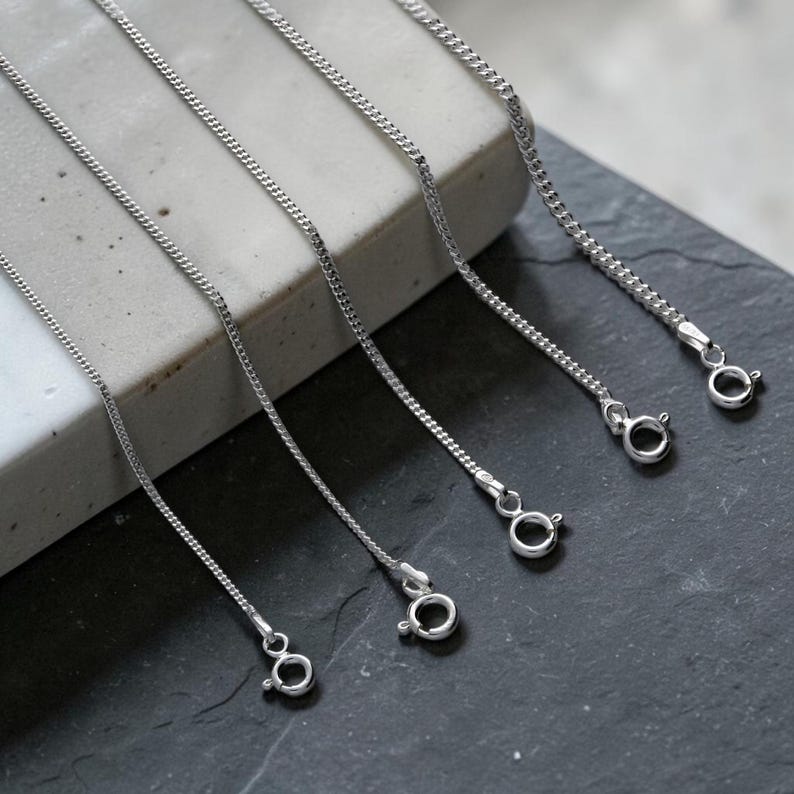 May include: Close-up of several silver-colored chain necklaces with lobster claw clasps. The chains are displayed on a gray and black surface. The chains have a delicate, textured design.
