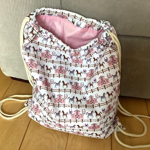 May include: A white drawstring bag featuring a repeating pattern of horses, barns, and horseshoes in brown and pink. The interior lining is pink, and the drawstring cords are cream-coloured.