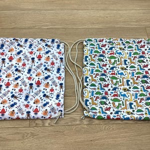 May include: Two drawstring bags. One has a pattern of robots in red, blue, and orange on a white background. The other has a pattern of colourful dinosaurs on a white background. Both have beige drawstrings.