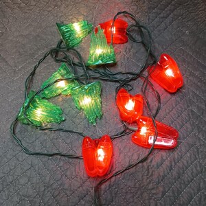 Könnte beinhalten: Eine dekorative Lichterkette mit durchscheinenden grünen schlossförmigen Abdeckungen und durchscheinenden roten schuhförmigen Abdeckungen. Die Lichter sind durch ein dunkelgrünes Kabel verbunden und strahlen ein warmes Licht aus.
