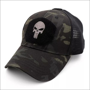 May include: A baseball cap featuring a dark green and black camouflage pattern with a black mesh back. The front panel displays a black rectangular patch with an embroidered light gray skull design centered on the crown of the hat.