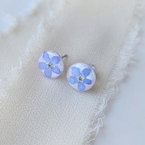 Puede incluir: Un par de pendientes de plata con una flor de nomeolvides prensada en cada uno. Las flores son de color azul claro con un centro amarillo.