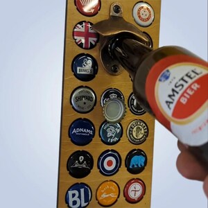 May include: A wooden wall-mounted bottle opener with a variety of bottle caps displayed. The bottle opener is bronze coloured and has a bottle cap attached to it. The bottle caps are arranged in a grid pattern and are from different brands of beer. A bottle of Amstel Bier is being opened with the bottle opener.