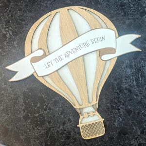 May include: A wooden hot air balloon with a banner that reads "Let the adventure begin". The balloon is white with brown stripes and has a brown wicker basket.
