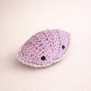 Può includere: Un piccolo peluche all'uncinetto a forma di mezzaluna di colore lavanda tenue. L'oggetto presenta un motivo a coste testurizzato e un bordo all'uncinetto beige chiaro lungo il perimetro. Due piccole perline nere fungono da occhi.