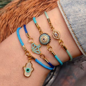 May include: Four gold-toned bracelets with blue and turquoise accents. One bracelet features a Hamsa hand charm, another a turquoise design, and two others have round and oval jewel details. The bracelets are on a blue and green cord.