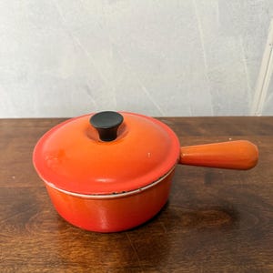 May include: A bright orange enamelled cast iron saucepan with a matching lid and a single long handle. The lid features a small black knob in the centre. The exterior of the pot and lid shows a vibrant gradient finish with minor wear along the rim.