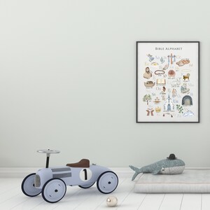May include: A framed print with a white background and colorful illustrations of Bible-themed alphabet letters. The print is titled "Bible Alphabet". A blue toy car and a gray whale toy are in the foreground.