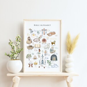 May include: A watercolor illustration of a Bible alphabet poster with images and words for each letter. The poster is framed and hanging on a wall.