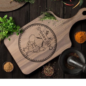 May include: A wooden chopping board with a handle, featuring a circular design with the text "Stevens BBQ" and a diagram of a chicken with different cuts labelled. The board is surrounded by various spices and herbs.