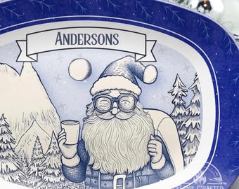 May include: A blue oval serving platter with a winter scene featuring a cartoon Santa Claus wearing glasses and holding a cup. The platter has a white banner with the text "Andersons" on it. The background is a blue and white starry sky with a mountain range and pine trees.