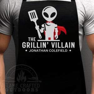 Puede incluir: Delantal negro con un extraterrestre blanco con una capa roja sosteniendo una espátula. El texto dice "The Grillin' Villain - Jonathan Colefield".