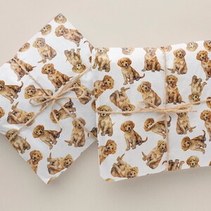 Puede incluir: Dos hojas de papel de regalo blanco con un patrón repetido de cachorros golden retriever. Las hojas están atadas con cuerda.