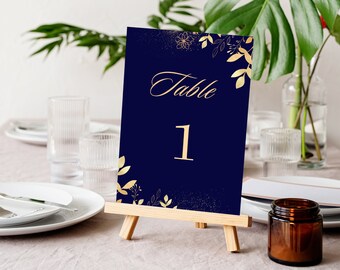 May include: A navy blue table number card with gold foil lettering and floral accents. The card reads "Table 1" and is displayed on a wooden easel.