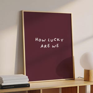 May include: A framed art print with a burgundy background and the white text "HOW LUCKY ARE WE." The frame is gold-colored. The print is displayed on a wooden shelf with a white globe lamp and a stack of books.