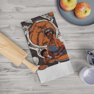 Op de afbeelding: Een witte keukendoek met een kleurrijke illustratie van een vrouw en een kind die taarten bakken. De illustratie bevat een grote kom met wortelen en een deegroller. Twee appels liggen op een blauw bord.
