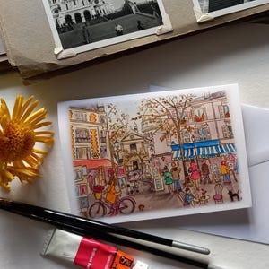 Puede incluir: Una pintura de acuarela de una escena callejera parisina con un caf&eacute;, &aacute;rboles y personas. El caf&eacute; tiene un toldo azul y est&aacute; decorado con flores. Los &aacute;rboles son verdes y tienen hojas amarillas. Las personas visten ropa colorida y caminan, se sientan y hablan.