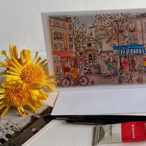 Puede incluir: Una pintura de acuarela de una escena callejera parisina con gente caminando y comprando. La pintura est&aacute; en una tarjeta blanca con un sobre blanco. Hay un tubo de pintura de acuarela en primer plano.