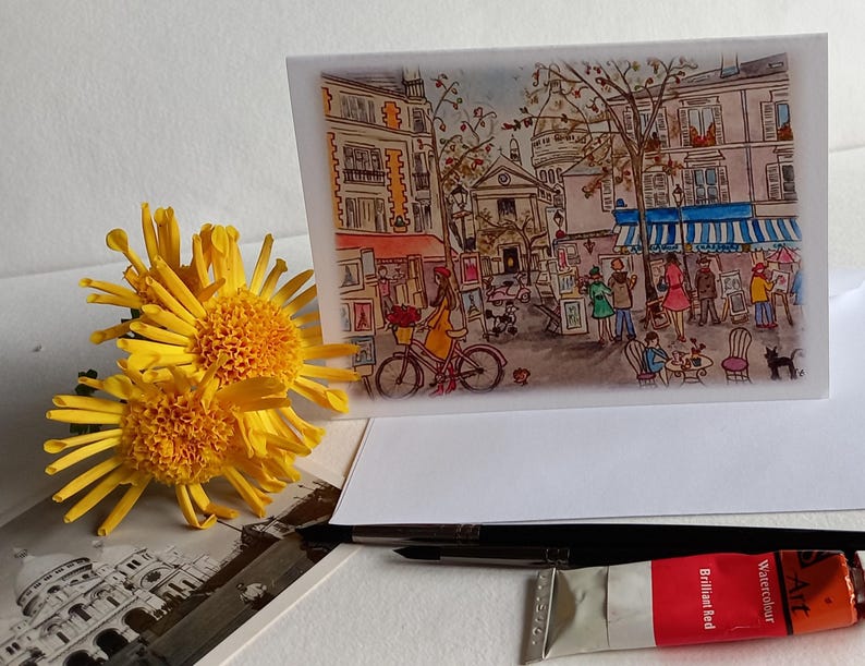 Puede incluir: Una pintura de acuarela de una escena callejera parisina con gente caminando y comprando. La pintura est&aacute; en una tarjeta blanca con un sobre blanco. Hay un tubo de pintura de acuarela en primer plano.