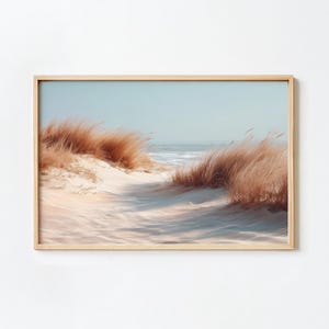 Könnte beinhalten: Ein gerahmter Druck einer Küstenlandschaft. Das Kunstwerk zeigt einen sandigen Pfad, der zum Meer führt, flankiert von hohen, goldbraunen Gräsern. Der Himmel ist hellblau, was eine ruhige und natürliche Szene schafft.