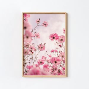 Può includere: Una stampa artistica da parete rettangolare in una cornice di legno chiaro che mostra una vista dal basso di numerosi fiori di cosmo rosa che si protendono verso un cielo tenue, pallido e nuvoloso. La composizione cattura petali delicati e steli.