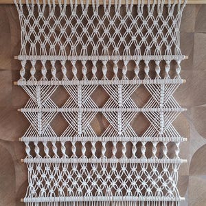 May include: Cream-colored macrame wall hanging with a geometric design. The piece features diamond and knot patterns, with wooden dowels at intervals. The bottom edge has a fringe. A decorative textile for home decor.