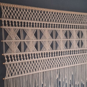 Könnte beinhalten: Cremefarbener Makramee-Wandbehang mit geometrischen Mustern und Fransen. Das Design zeigt Diamant- und X-förmige Knoten, die an einem natürlichen Holzstab aufgehängt sind. Ein dekoratives Stück für die Inneneinrichtung.