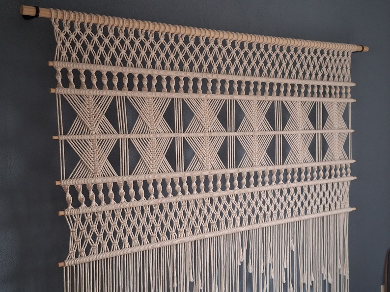 May include: Cream-colored macrame wall hanging with geometric patterns and fringe. The design features diamond and X-shaped knots, suspended from a natural wooden dowel. A decorative piece for home decor.