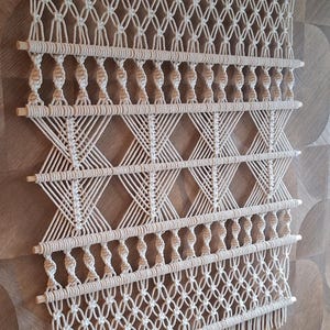 May include: Cream-colored macrame wall hanging with a geometric design. The piece features diamond and crisscross patterns, with wooden dowels at the top and bottom. The bottom edge has a fringe detail. A decorative textile for home decor.