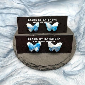 May include: Two pairs of blue and white butterfly earrings with glitter accents. The earrings are displayed on black cards with the text "Beads by Batsheva Handmade Jewelry".