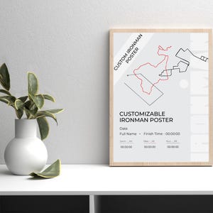 May include: A framed poster with a light wood finish. The poster features a red and black graphic design of a race course, with the text "CUSTOM IRONMAN POSTER" and "CUSTOMIZABLE IRONMAN POSTER". The poster includes fields for date, name, and finish time.