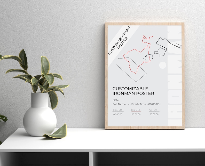 May include: A framed poster with a light wood finish. The poster features a red and black graphic design of a race course, with the text "CUSTOM IRONMAN POSTER" and "CUSTOMIZABLE IRONMAN POSTER". The poster includes fields for date, name, and finish time.