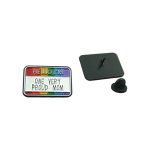 Pode incluir: Um broche de esmalte arco-íris com o texto "HELLO, I'M ONE VERY PROUD MOM" em um fundo branco. O broche tem um verso de metal preto e um pino de segurança.