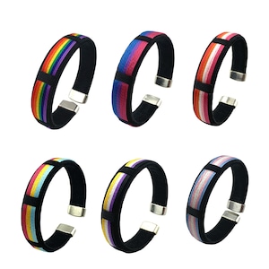 Könnte beinhalten: Sechs bunte Armbänder mit verschiedenen Pride-Flaggen-Designs. Jedes Armband besteht aus schwarzem Stoff mit einem bunten, gewebten Streifenmuster. Die Armbänder sind verstellbar und haben einen Magnetverschluss.