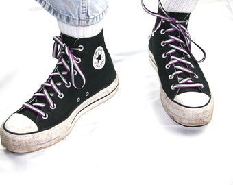 May include: A pair of black high-top Converse sneakers with white laces and purple and white striped laces. The laces are tied in a criss-cross pattern.