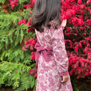 May include: A pink floral dress with long sleeves and a matching pink sash tied at the waist. The dress has a vintage-inspired print and is worn by a child standing in front of a red bush.