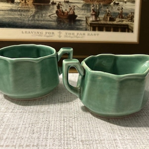 May include: A set of two sage green ceramic vessels, consisting of a sugar bowl with two side handles and a creamer pitcher with a single handle and a pouring spout. Both pieces feature a scalloped rim and a smooth, glossy finish.