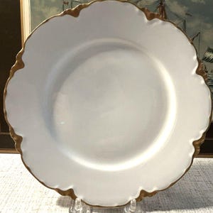 May include: A round white ceramic dinner plate featuring a scalloped edge with a metallic gold trim. The plate has a smooth, glossy finish and a wide, flat center area, presented on a clear plastic stand for display purposes.