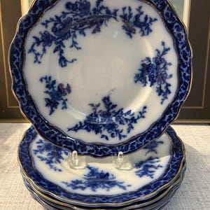 May include: A stack of white ceramic plates featuring a deep blue floral pattern and a scalloped edge with a gold trim. The plates have a wide, dark blue border decorated with intricate scrolling designs and a central white area with floral motifs.