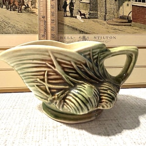 May include: A ceramic creamer pitcher featuring a molded shell and leaf design with a glossy green and brown glaze. A wooden ruler is positioned behind the pitcher, showing a height of approximately 3 inches and a width of about 5 inches.