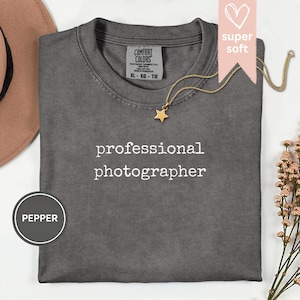 May include: A grey Comfort Colors t-shirt with the words "professional photographer" in white. A gold star pendant necklace and a pink ribbon with the words "super soft" are on the shirt. A brown hat and flowers are in the background.
