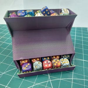 May include: A purple dice box filled with various colorful dice. The dice are in different colors, including blue, red, yellow, and purple, with white or yellow dots. The box is open, revealing the dice inside.